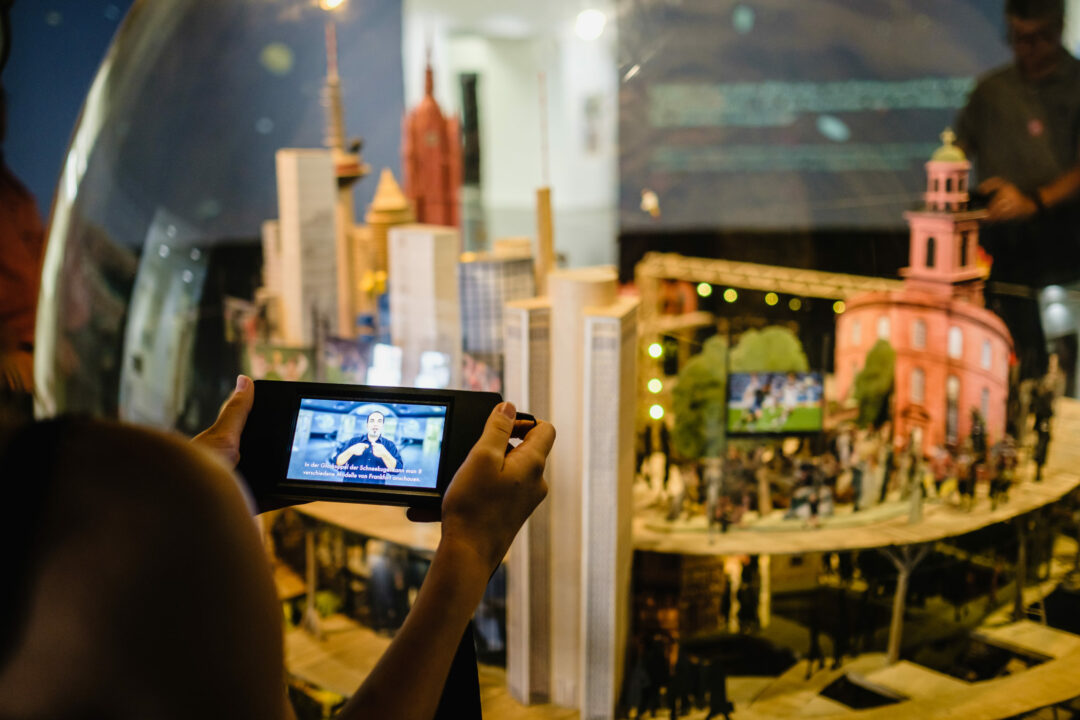 Auf einem digitalen Endgerät wird vor einem Ausstellungsexponat ein Film abgespielt.