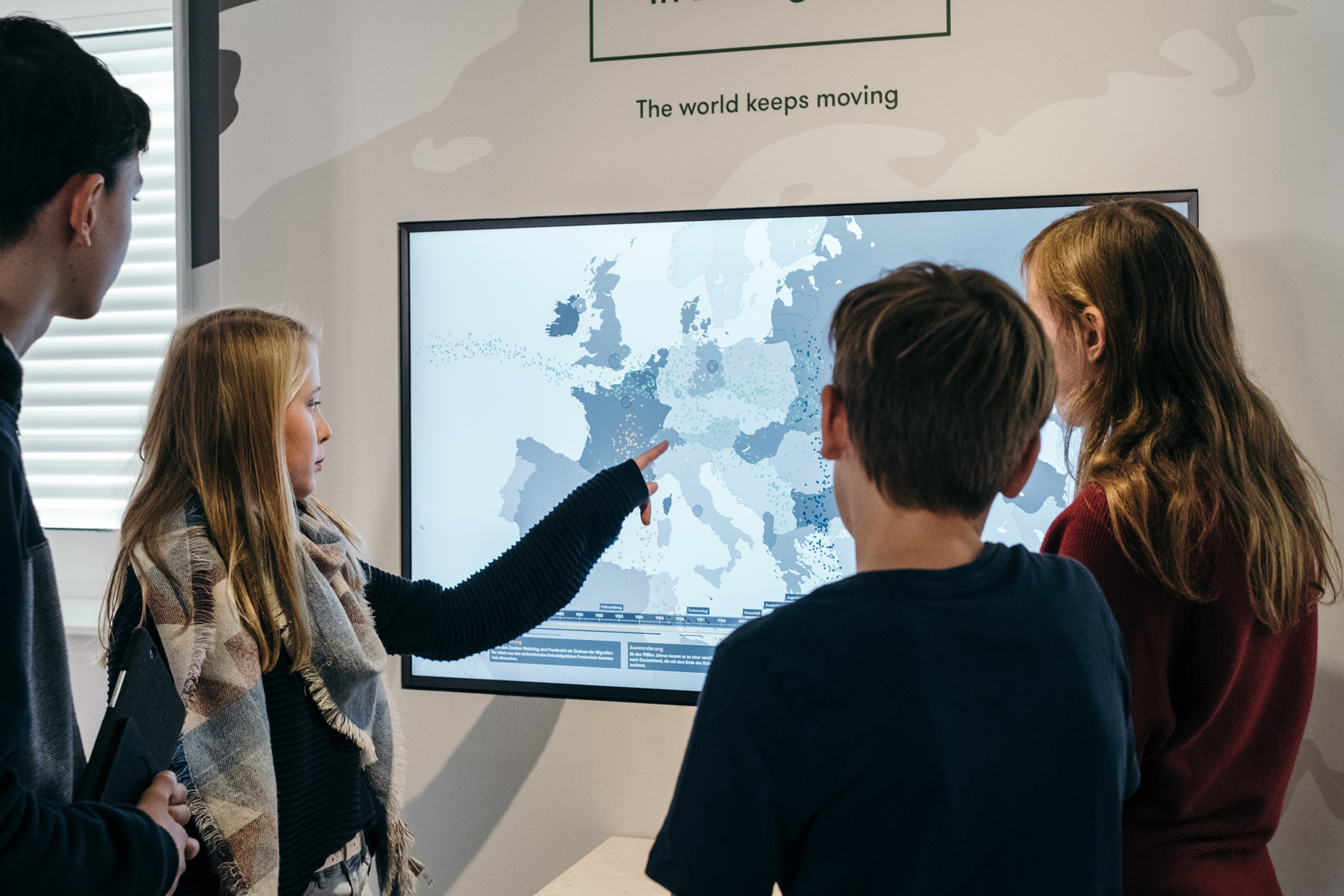 4 Jugendliche Stehen vor einem Bildschirm mit einer Weltkarte, über dem Bildschirm stehen die Worte: "The world keeps moving".