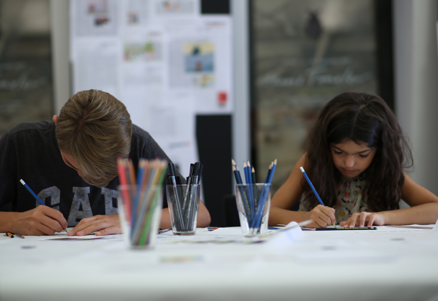 zwei Kinder, die an einem Tisch sitzen und zeichnen. In der Mitte des Tisches befinden sich drei Gläser mit Stiften.